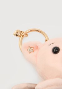 Porte-clés en peluche rose avec œil en perle noire, détail en strass en forme d'étoile dorée sur l'oreille, et anneau doré avec un petit pendentif en forme de cœur gravé.