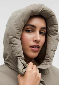 Padded béžová bunda s kapucňou a plyšovou textúrou. Kapucňa je nadmerná a zhrnutá, obsahuje zips a gombíkový uzáver.