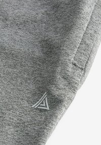 Gros plan sur un tissu gris à motif texturé, avec une poche cousue et un petit logo triangulaire en gris clair.