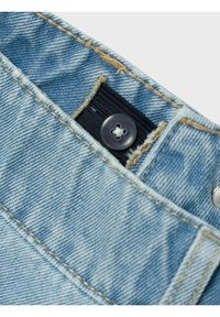 Tejido de mezclilla de color azul claro con costuras doradas, que presenta una banda elástica negra y un botón gris en la cintura. Vista en primer plano de los detalles.