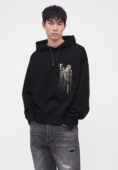 Jeune homme portant un sweat à capuche noir Emporio Armani et un jean gris délavé et déchiré, debout les mains dans les poches devant un fond uni.