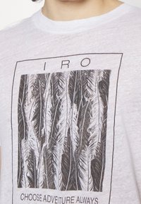 Biała bawełniana koszulka z grafiką czarno-białego pióra oraz napisem "IRO" i "W ZAWSZE WYBIERAJ PRZYGODĘ" w prostokątnej ramce.