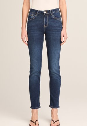 Slim-fit dark blue denim jeans worn with black flip-flops on a person standing against a beige background.