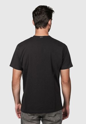 Hombre con cabello oscuro que lleva una camiseta negra lisa de manga corta y pantalones vaqueros grises, de espaldas sobre un fondo claro y liso.