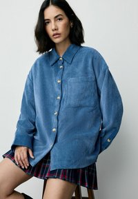 Camicia di velluto a coste blu con abbottonatura frontale, colletto alla francese, taschino sul petto e maniche lunghe, abbinata a una gonna a quadri.