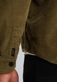 Olijfgroene corduroy jacket met verticale ribbelstructuur, voorzien van een knoopsluiting aan de manchetten, logo-opschrift en zwarte accenten. Geschikt voor verschillende gelegenheden.