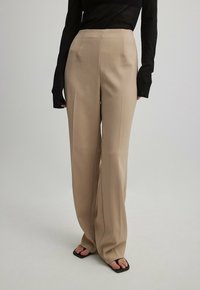 Pantalons beige à larges jambes avec une texture lisse, dotés de plis sur le devant et d'une taille haute. Associés à un haut noir à manches longues et des sandales noires.