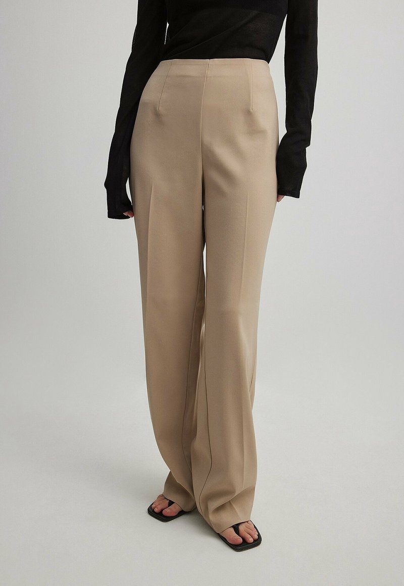 Pantalons beige à larges jambes avec une texture lisse, dotés de plis sur le devant et d'une taille haute. Associés à un haut noir à manches longues et des sandales noires.