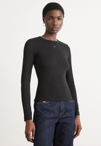Black long-sleeve ribbed top with a round neckline and logo detail; paired with dark blue denim jeans. Simple and fitted design.