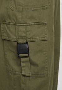 Olive green cargo pant section featuring a patch pocket with a black plastic buckle and reinforced stitching, highlighting its durable material.