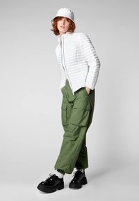 Giacca bianca a zip con effetto piumato e righe orizzontali, abbinata a pantaloni cargo verdi larghi. Le scarpe platform nere completano il look.