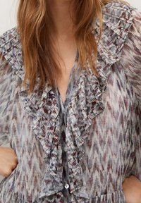 Nainen, joka on pukeutunut puoliksi läpikuultavaan kuvioituun puseroon, jossa on röyhelökaulus ja etuosa, sävyissä harmaa, violetti ja ruskea.