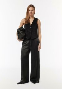 Black sleeveless vest with a v-neck and three buttons, paired with wide-leg black trousers. The model holds a black, soft-textured bag.