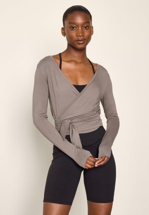Ung kvinde iført en taupe omslagstop med tommelhuller over en sort sports-bh og stramme sorte cykelshorts, stående mod en ensfarvet baggrund.