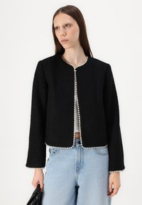 Veste noire texturée avec un col rond et des accents en perles sur le devant et les manches, associée à un jean en denim bleu clair.