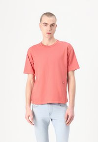 Kortermet korall T-skjorte i myk bomull med rund hals og et lite merke på nederste søm. Kombinert med lyseblå jeans.