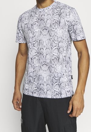 Homme portant un t-shirt manches courtes blanc et noir à imprimé serpent avec un pantalon noir sur un fond uni.