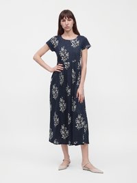 Femme portant une robe midi bleu marine à imprimés floraux blancs, manches courtes, et des ballerines beiges pointues, debout avec une main sur la hanche.