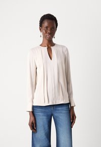 Lys beige bluse med V-hals, plisser foran og lange ermer. Stoffet ser glatt ut og faller mykt. Bæres med blå jeans.