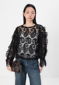 Schwarze Spitzenbluse mit Paisleymuster, durchsichtigen Abschnitten, gerafften langen Ärmeln und einem runden Ausschnitt, kombiniert mit blauen Jeans und einer dunklen Handtasche.