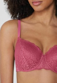 Różowy stanik koronkowy z wyprofilowanymi miseczkami, falbanastymi krawędziami i regulowanymi ramiączkami. Posiada szczegółowe wzory kwiatowe i gładką teksturę.