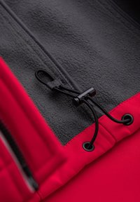 Cordon noir avec toggle sur l'ourlet de la veste rouge au-dessus de la doublure polaire grise avec fermeture éclair noire visible sur le côté.