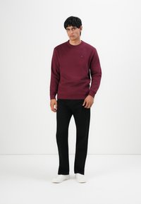 Hombre de pie contra un fondo blanco, vistiendo un sudadera burdeos, pantalones negros y zapatillas blancas, mirando hacia adelante.