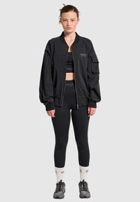 Czarna kurtka bomberka z zamkiem z przodu, ściągaczami przy mankietach i bocznymi kieszeniami. Noszona na crop top i legginsy. Szare sneakersy z teksturowaną podeszwą.