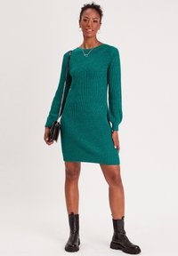 Vestido de punto acanalado en color verde azulado con mangas largas, escote en forma de V y silueta ajustada, combinado con botas negras hasta el tobillo.