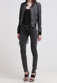 Donna che indossa una giacca di pelle nera, una maglia nera, jeans skinny effetto stone-washed e sandali neri con tacco aperti, in piedi davanti a uno sfondo semplice.