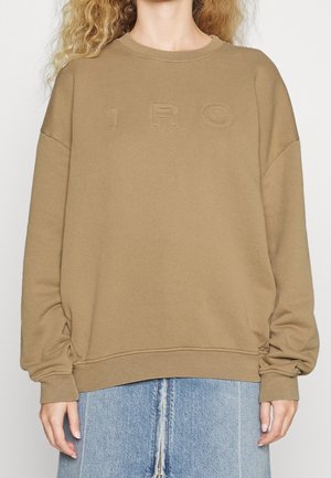 Beiger Oversize-Sweatshirt mit geprägten "IRO"-Buchstaben, getragen mit einem hellblauen Jeansrock mit Frontreißverschluss.
