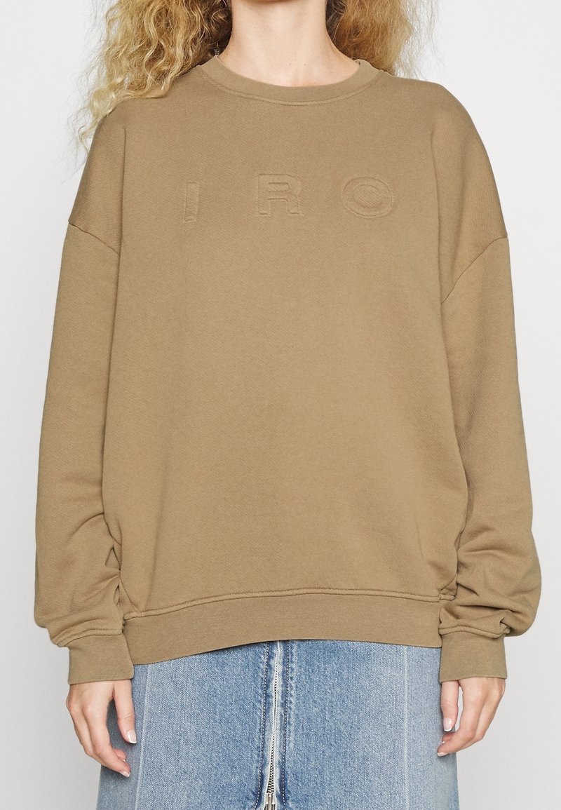 Sweat-shirt oversize beige avec des lettres "IRO" en relief, porté avec une jupe en jean bleu clair dotée d'une fermeture éclair à l'avant.