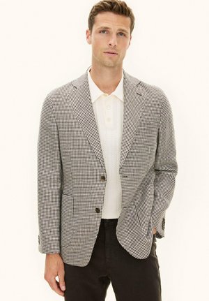 Mężczyzna w brązowo-białym blezerze w jodełkę, białej dzianinowej koszulce polo i czarnych spodniach, stojący z jedną ręką w kieszeni na jednolitym tle.