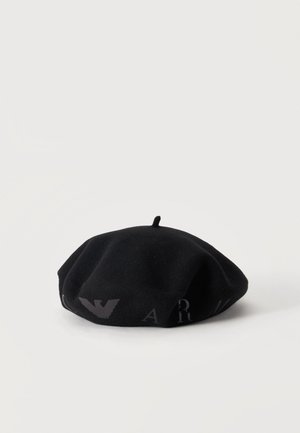 Béret en laine noir avec petite tige sur le dessus et logo sombre subtil ainsi que des lettres sur le côté, posé sur fond blanc.
