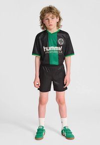Schwarz-grünes Sportoutfit mit horizontalen Streifen, kurzen Ärmeln und Shorts. Das Shirt hat ein Logo und grüne Akzente. Grüne Turnschuhe vervollständigen das Outfit.