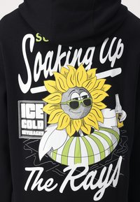 Felpa nera con un vivace grafico di un girasole giallo che indossa occhiali da sole verdi, mentre tiene una bottiglia, con il testo "Assaporando i raggi."