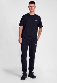 Černé bavlněné tričko a tepláky s elastickými manžetami, s malým logem na hrudi a nohavici. Hladká textura a volný střih.