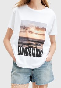 Bílá bavlněná trička s grafickým potiskem západu slunce nad oceánem, se slovy "ALLSAINTS" v tučných černých písmenkách na spodní části.
