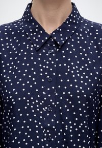 Μπλε ναυτικό πουκάμισο με λευκές πουά βούλες, με μυτερό γιακά και τέσσερα εμφανή κουμπιά.