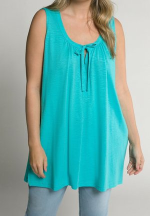 Vrouw draagt een mouwloze turquoise tuniek met een strikdetail aan de voorkant en lichtblauwe spijkerbroek, staand tegen een effen achtergrond.