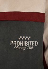 Tissu vert foncé et beige avec une bande horizontale rouge et un texte brodé « PROHIBITED Racing Club » sous un motif de drapeau à damier.