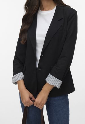 Vrouw draagt zwarte blazer met opgerolde gestreepte manchetten, wit T-shirt, blauwe spijkerbroek, houdt bruine tasgrepen vast voor een witte achtergrond.