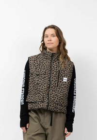 Leopardprint vest in beige en zwarte stof met een hoge kraag, ritssluiting en twee voorkruiszakken. Gemaakt over een zwart shirt met lange mouwen.