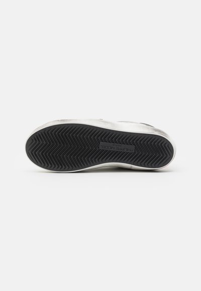 Black rubber sole with zigzag pattern; white side with subtle texture; branding visible on the sole.