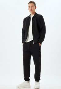 Schwarze Zip-Jacke über einem weißen T-Shirt, kombiniert mit schwarzen Jogginghosen und weißen Turnschuhen. Weicher Stoff, bequemer Schnitt und elastische Bündchen.