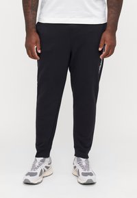 Schwarze sportliche Jogginghosen aus glattem Stoff, mit tapered Fit, seitlichen Taschen und weißer Logo-Detailierung entlang des linken Beins. Kombiniert mit grauen Sneakers.