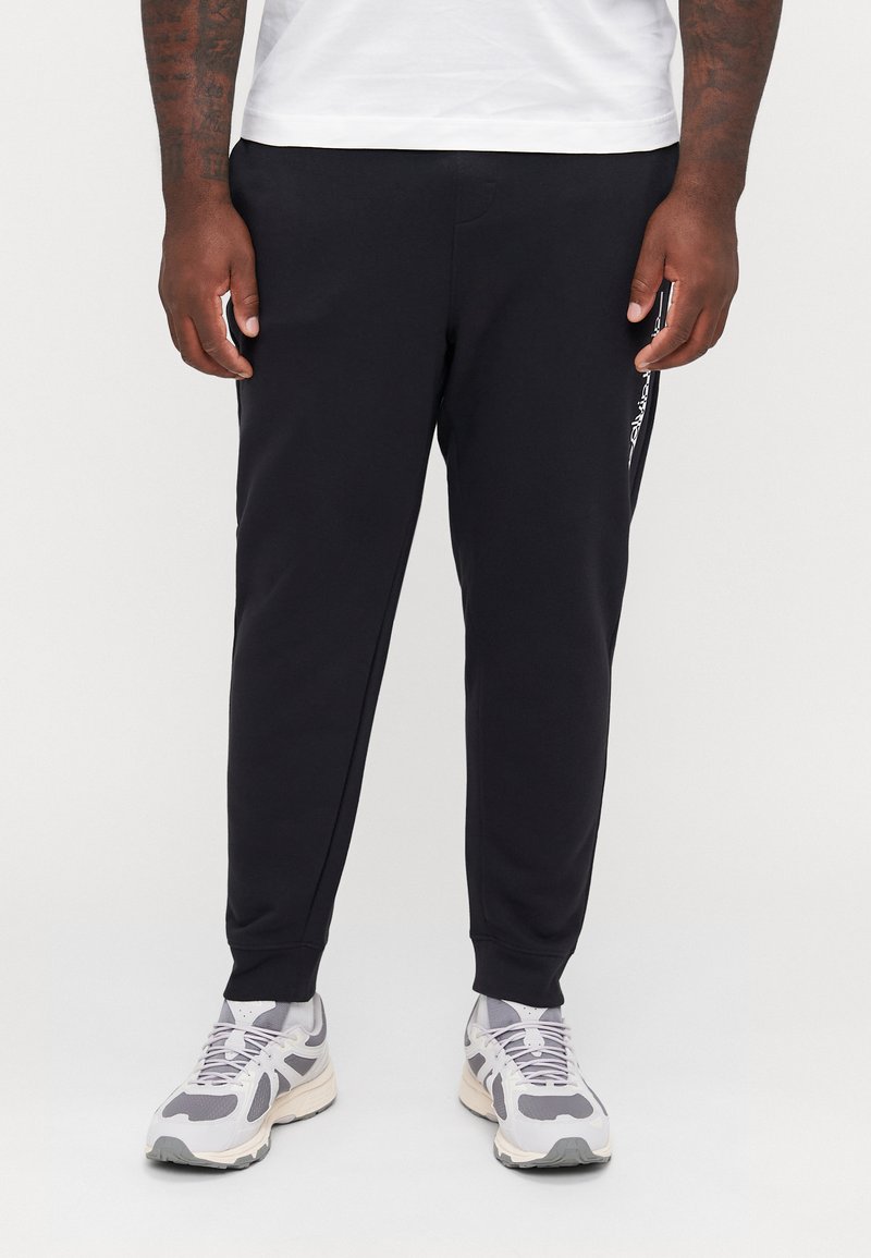 Schwarze sportliche Jogginghosen aus glattem Stoff, mit tapered Fit, seitlichen Taschen und weißer Logo-Detailierung entlang des linken Beins. Kombiniert mit grauen Sneakers.