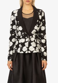Schwarzer und weißer Blumenblazer mit Spitzenstruktur, mit Reverskragen, Ein-Knopf-Verschluss und zwei vorderen Taschen, über einem schwarzen Kleid getragen.