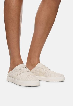 Beige slip-on shoes with textured fabric featuring a geometric pattern, rounded toe, and cushioned white sole. Two adjustable straps on top.