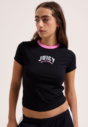 Junge Frau mit langen braunen Haaren, die ein schwarzes T-Shirt mit rosa Kragen und dem Schriftzug "JUICY Est. Los Angeles" trägt, schaut seitlich.